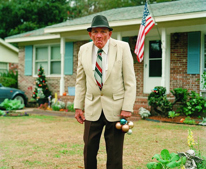 Photograph of an older man in dapper attire outside a ranch-style house